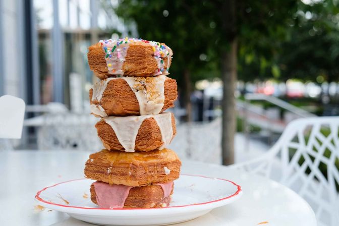 Stack of flaky cronuts with pink, white, and caramel glazes and rainbow sprinkles, served on a white plate at an outdoor café table