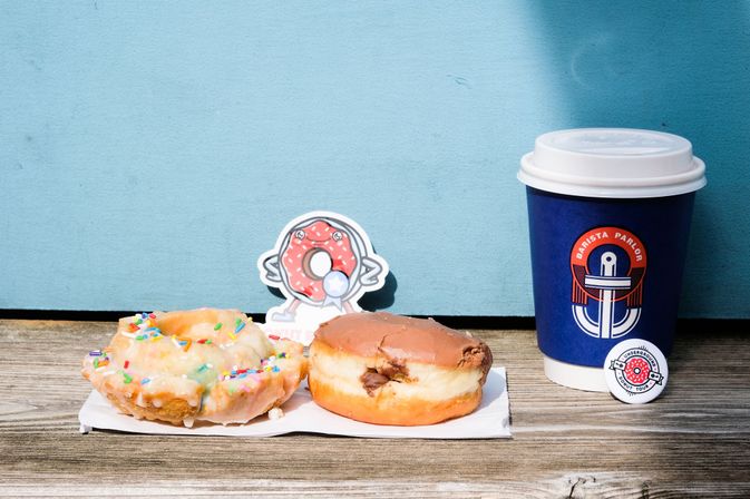 Two donuts—a glazed ring with rainbow sprinkles and a chocolate-filled round—on a napkin beside a blue paper coffee cup with white lid on a weathered wooden bench in front of a light blue wall, with a playful donut character cutout behind them.