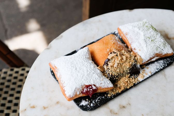 Three golden, powdered-sugar beignets on a black slate with jam and chocolate fillings and crushed nuts, served on a marble café table