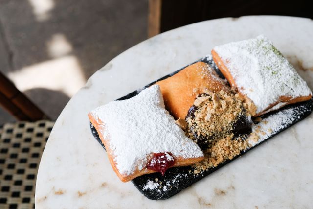 Three golden, powdered-sugar beignets on a black slate with jam and chocolate fillings and crushed nuts, served on a marble café table