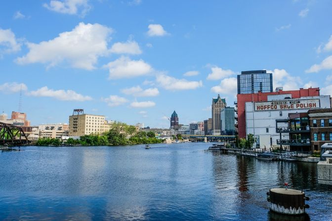 Riverfront city skyline under a bright blue sky, calm water with a small boat, and a brick building featuring a faded vintage shoe-polish advertisement along the waterfront.