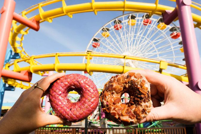 Two hands hold donuts—one pink glazed, one topped with crispy crumble—framed by a bright yellow roller coaster and colorful Ferris wheel on a sunny amusement park day.