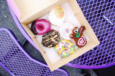 Box of six colorful gourmet donuts — assorted glazes, sprinkles, cereal and chocolate toppings plus a character-shaped donut — sitting on a purple metal outdoor café table