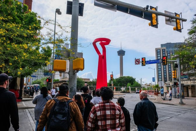 Downtown San Antonio intersection with pedestrians waiting at a crosswalk, a bright red abstract sculpture and the Tower of the Americas in the background