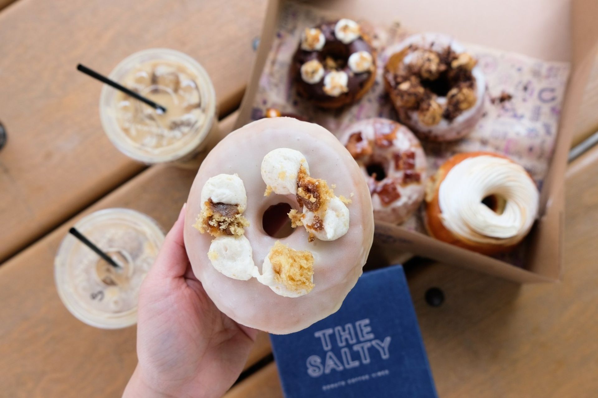 Top-down view of a hand holding a glazed artisan donut topped with cream dollops and crumbly streusel, with a box of assorted donuts and two iced coffees on a wooden picnic table.
