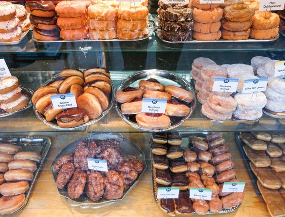 Donut shop glass display packed with assorted doughnuts — glazed rings, powdered sugar stacks, chocolate‑topped bars, filled Bavarian and raspberry donuts, and apple fritters.