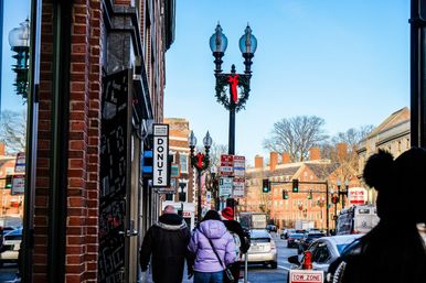 Downtown city street with brick buildings, pedestrians in winter coats, holiday wreaths on lampposts, a vertical "donuts" shop sign, traffic lights and cars under a clear blue sky.