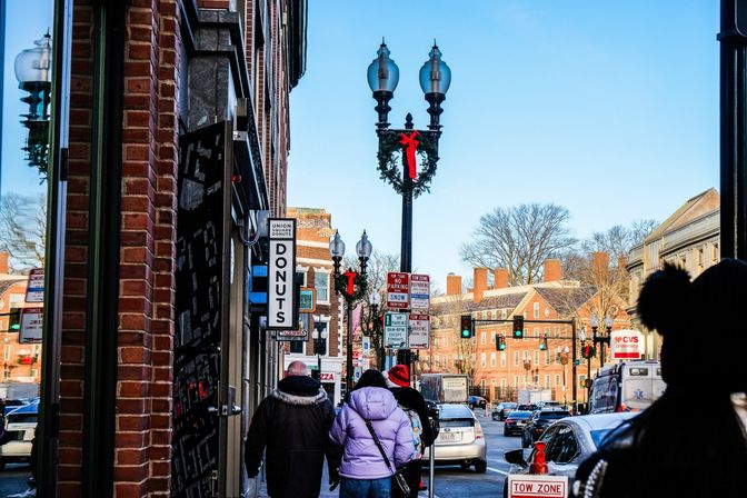 Downtown city street with brick buildings, pedestrians in winter coats, holiday wreaths on lampposts, a vertical "donuts" shop sign, traffic lights and cars under a clear blue sky.