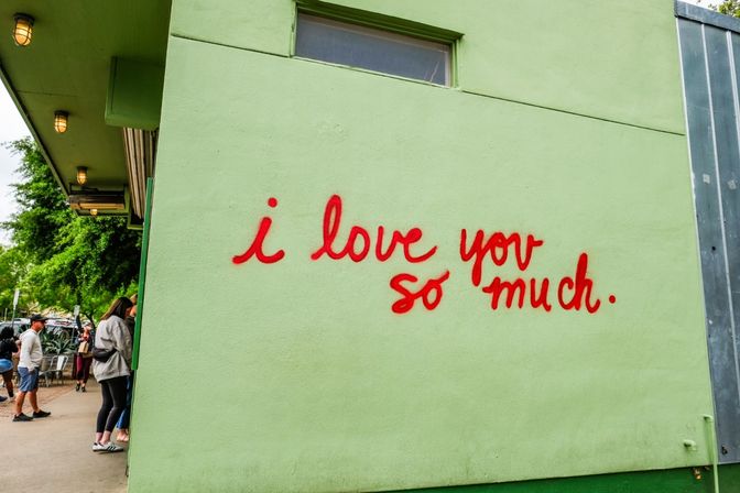 Red handwritten mural reading "i love you so much." on a pale green exterior wall above an urban sidewalk with people under an awning and leafy street trees.