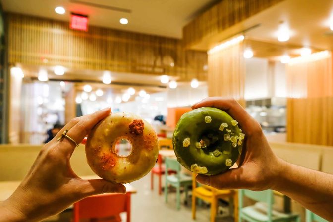 Two hands hold colorful artisan donuts—white glazed with red crumbs and green matcha topped with candied pieces—inside a modern wood-accented cafe with colorful chairs