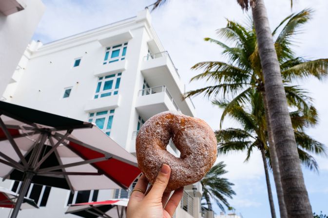 Hand holding a powdered-sugar donut against a white beachfront building, palm trees, outdoor café umbrellas and blue sky.