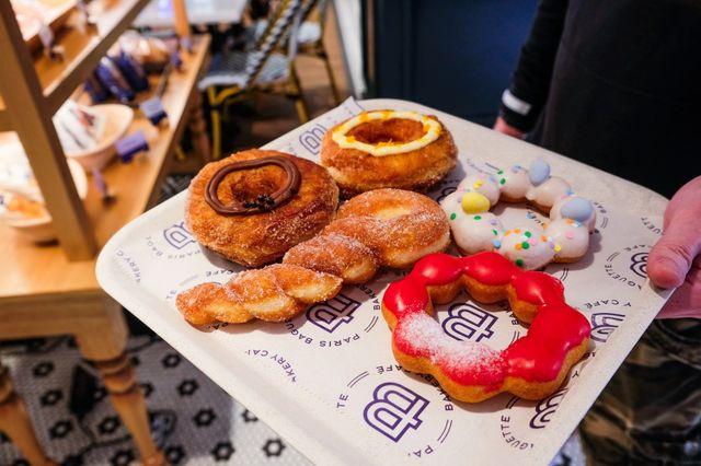 Assorted colorful donuts and sugar-coated crullers on a paper-lined tray at a bakery counter — chocolate, lemon, pink-frosted and sprinkle-topped pastries.