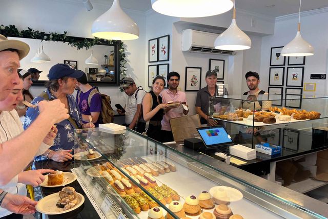 Busy neighborhood bakery interior with glass display case of éclairs, croissants and assorted pastries, customers lining the counter under modern pendant lights.