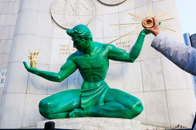 Green Spirit of Detroit statue at a marble downtown memorial holding a tiny gilded figure while a person off-frame offers a glazed donut to its outstretched hand.