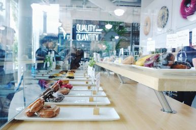 Assorted colorful artisanal donuts on white plates in a glass display case at a modern urban bakery cafe, with a neon sign reading "QUALITY over QUANTITY" glowing in the background.