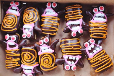 Box of chocolate‑coated rectangular pastries decorated with pink, orange and white icing as playful Halloween monster faces