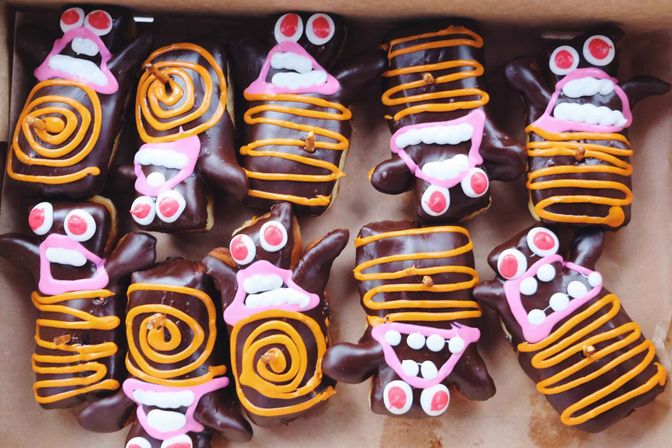 Box of chocolate‑coated rectangular pastries decorated with pink, orange and white icing as playful Halloween monster faces