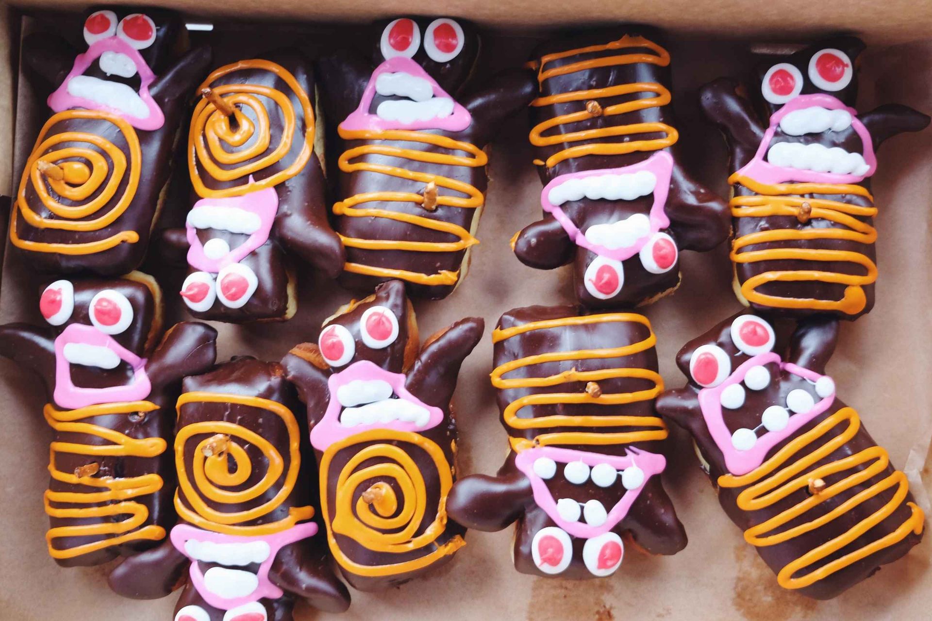 Box of chocolate‑coated rectangular pastries decorated with pink, orange and white icing as playful Halloween monster faces