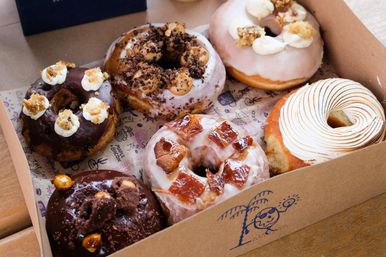 Box of assorted gourmet donuts in a takeaway box on a wooden table — chocolate-glazed, cream-topped, caramel and bruleed meringue varieties.