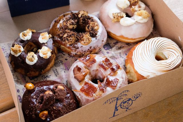 Box of assorted gourmet donuts in a takeaway box on a wooden table — chocolate-glazed, cream-topped, caramel and bruleed meringue varieties.