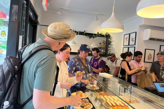 Busy neighborhood bakery interior with a line of customers selecting éclairs, cream-filled pastries and samples from a glass display case under white pendant lights