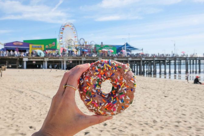 Hand holding a sprinkled glazed doughnut in foreground with a sunlit sandy beach, colorful amusement pier and Ferris wheel in the background
