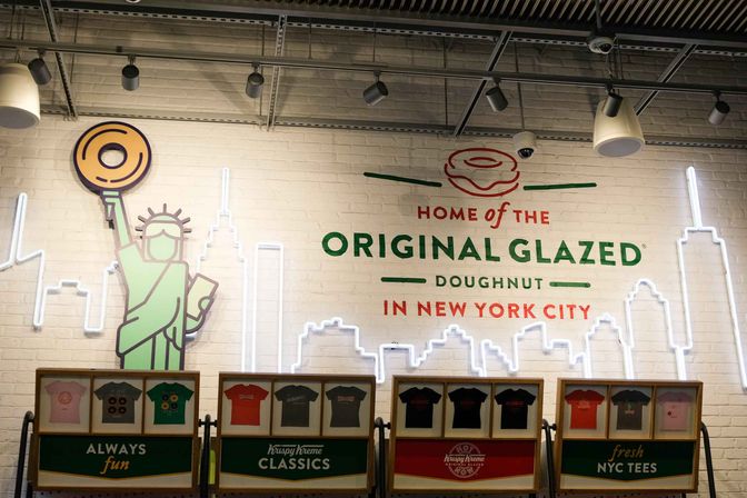 Bright New York City doughnut shop mural with a stylized Statue of Liberty holding a giant glazed doughnut, neon skyline outline, and racks of colorful NYC t-shirts.