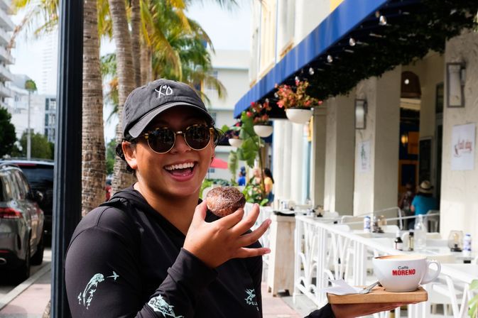Smiling person in a black cap and sunglasses holding a sugar-dusted pastry and a cup of coffee at a sunny palm-lined sidewalk café in a coastal city