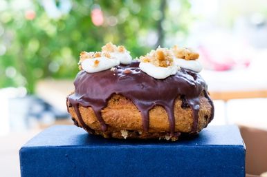 Chocolate‑glazed gourmet donut with whipped cream dollops and crumb topping, chocolate dripping over the edge, sitting on a blue box with a green outdoor bokeh background