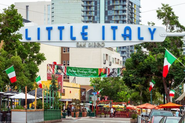 Arched Little Italy San Diego sign over a tree-lined street decorated with Italian flags, outdoor dining patios, orange umbrellas and downtown condos.
