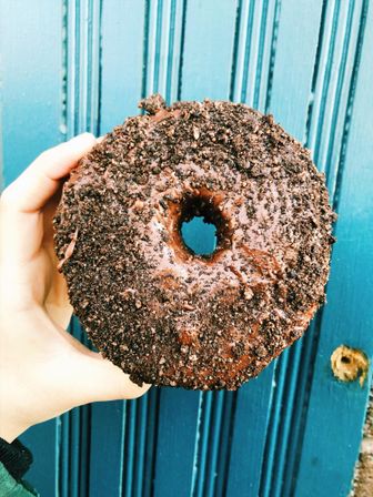 Hand holding a chocolate-glazed donut coated in crushed cookie crumbs against a teal-blue paneled door backdrop