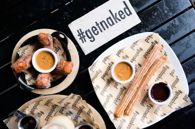 Top-down view of a dessert spread on a black café table: sugar-coated churros with caramel and chocolate dipping sauces, cinnamon-sugar fried dough bites with caramel dip, and a napkin stamped '#getnaked'.