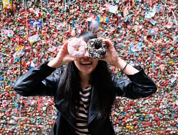 Smiling person in a leather jacket holding a pink sprinkled donut and a cookies-and-cream donut over their eyes in front of a colorful gum-covered wall.