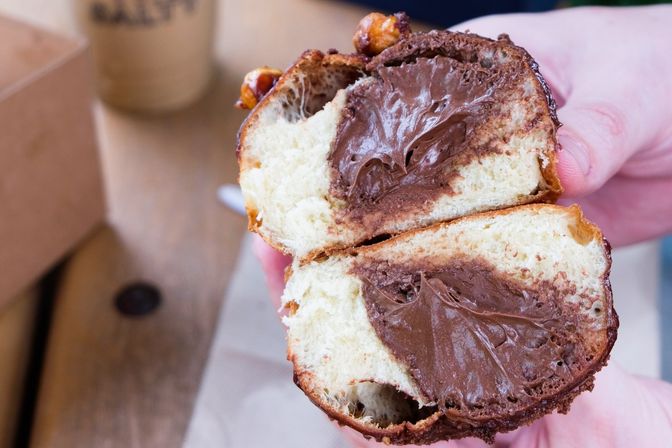 Close-up of a hand holding a split brioche bun filled with glossy chocolate-hazelnut spread, a handheld café pastry breakfast treat