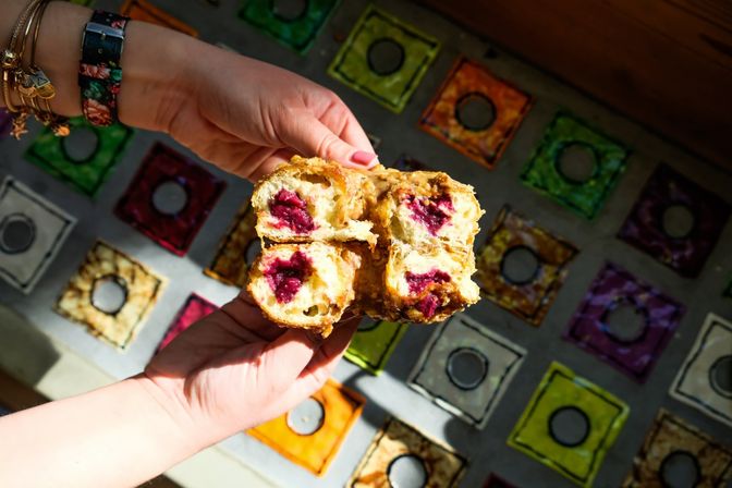 Close-up of hands tearing open a flaky pastry oozing vibrant berry filling over colorful square glass tiles.
