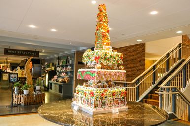 Tall multi-tiered gingerbread and candy display on a round marble table in a hotel lobby, decorated with icing, gumdrops, candy canes and tiny gingerbread houses next to a stairway