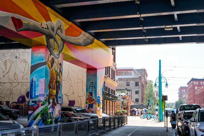 Colorful urban mural covering underpass pillars in a downtown street — painted cityscapes and figures beneath a dark overpass, parked cars, turquoise street lamps and pedestrians on a sunny block.