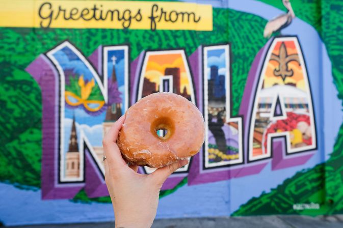 Glazed donut held in front of a colorful 'Greetings from NOLA' postcard-style mural in New Orleans