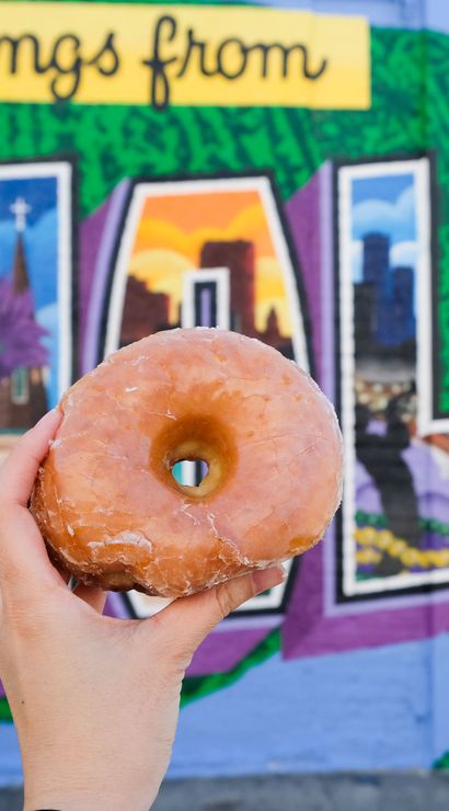 Glazed donut held in front of a colorful 'Greetings from NOLA' postcard-style mural in New Orleans