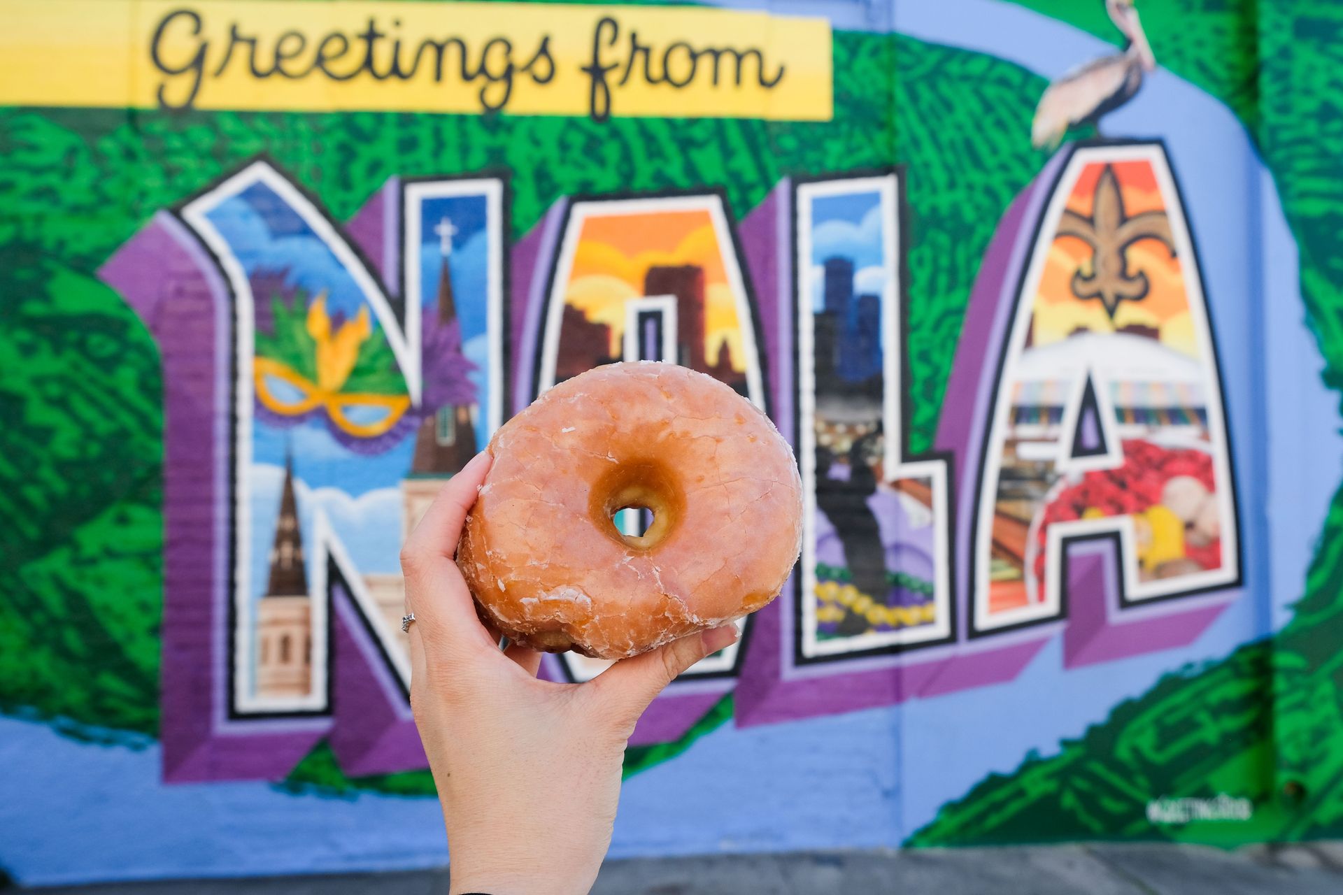 Glazed donut held in front of a colorful 'Greetings from NOLA' postcard-style mural in New Orleans