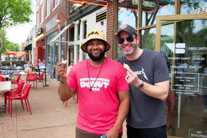 Two smiling men holding small donut bites on a lively downtown sidewalk outside a storefront, with red outdoor chairs, tables and trees lining the street.