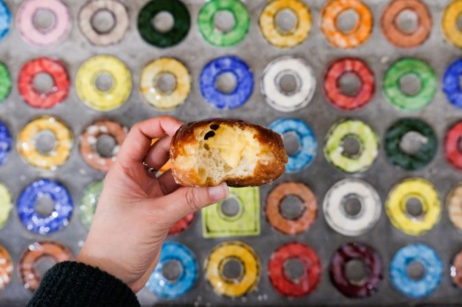 Hand holding a bitten custard-filled glazed donut against a vibrant wall of colorful glazed donut tiles.