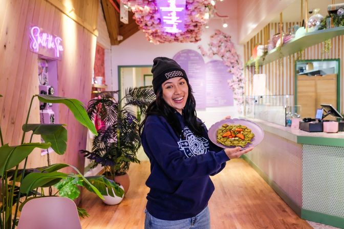 Smiling woman in a black beanie holding a plate of matcha waffles topped with sliced strawberries inside a trendy pastel pink and green cafe with neon sign, floral ceiling decor and potted plants.