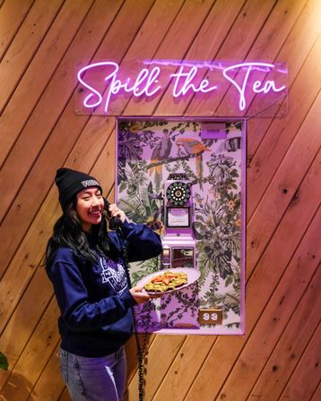 Smiling person in a beanie inside a wood-paneled cafe holding a plate of strawberry-topped waffles and speaking into a retro phone beneath a pink neon "Spill the Tea" sign.