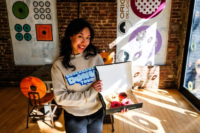 Smiling woman in a sunlit, brick-walled cafe holding an open box of colorful donuts with wooden chairs and circular mural art in the background.