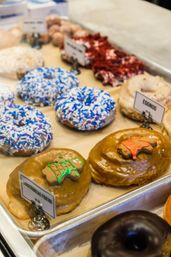 Assorted donuts in a bakery display case — caramel-glazed donuts topped with tiny gingerbread cookies, blue-and-white sprinkle donuts, an eggnog-labeled flavor, and a chocolate-glazed donut.