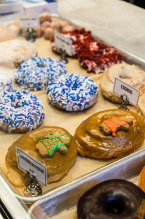 Assorted donuts in a bakery display case — caramel-glazed donuts topped with tiny gingerbread cookies, blue-and-white sprinkle donuts, an eggnog-labeled flavor, and a chocolate-glazed donut.