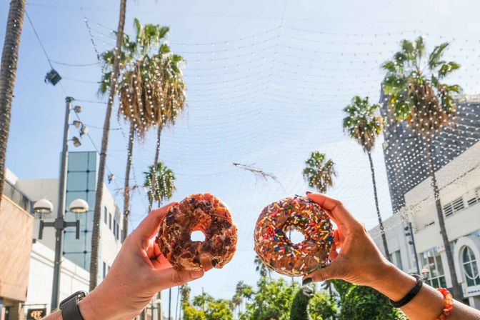 Two hands holding a colorful sprinkle-topped donut and a glazed bacon-topped donut against a sunny, palm-lined street with decorative hanging string lights
