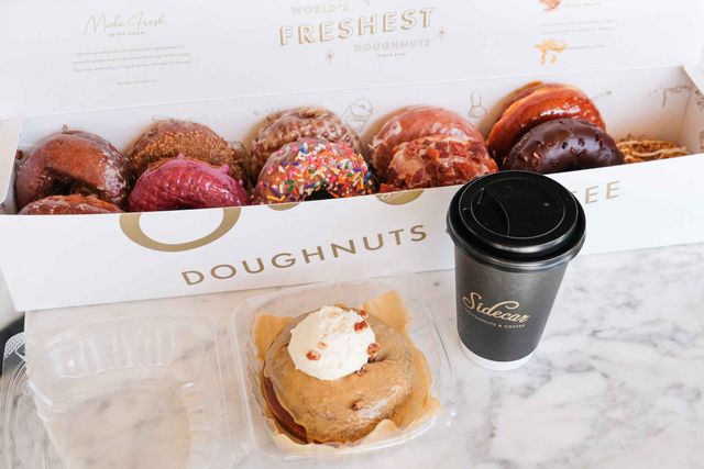 Assorted glazed and topped doughnuts in a bakery box — colorful sprinkle and chocolate varieties — plus a single cream-topped donut in a clear takeout container and a black to-go coffee cup on a marble countertop.