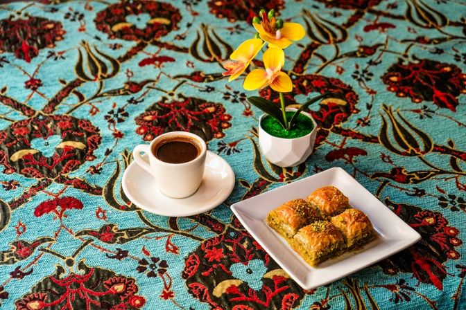 White cup of Turkish coffee on a saucer with a square plate of pistachio baklava and a small potted yellow orchid on a vibrant turquoise Ottoman-style patterned tablecloth.